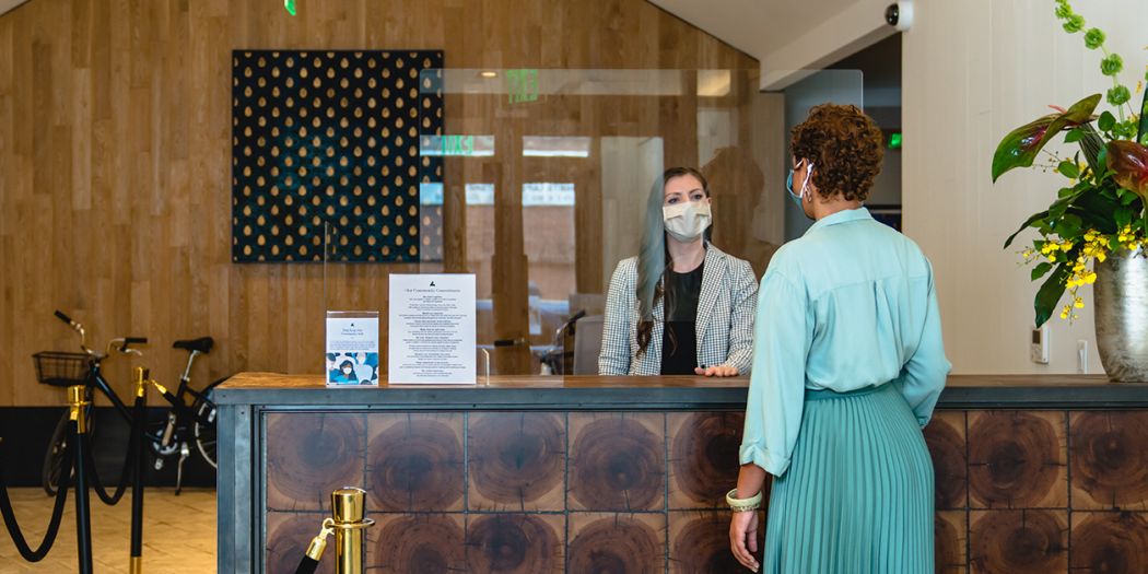 A woman wearing a face mask stands at the front desk, attending to a guest.