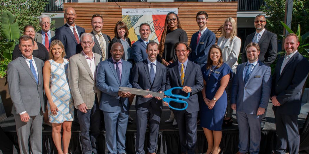 A group of Bozzuto business professionals posing in front of a building, ready to cut a ribbon with a big scissor.