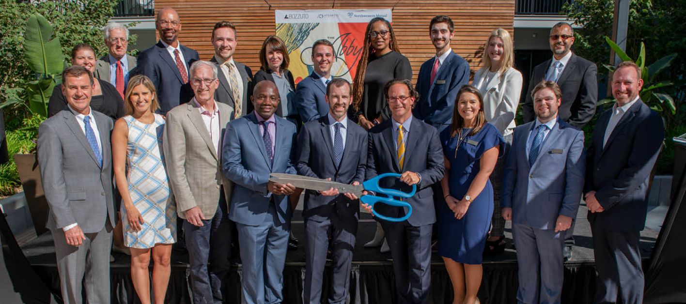 A group of Bozzuto business professionals posing in front of a building, ready to cut a ribbon with a big scissor.