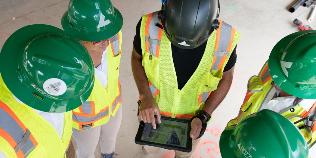 A Bozzuto group of construction workers in green vests & hard hats looking at a tablet, discussing plans.