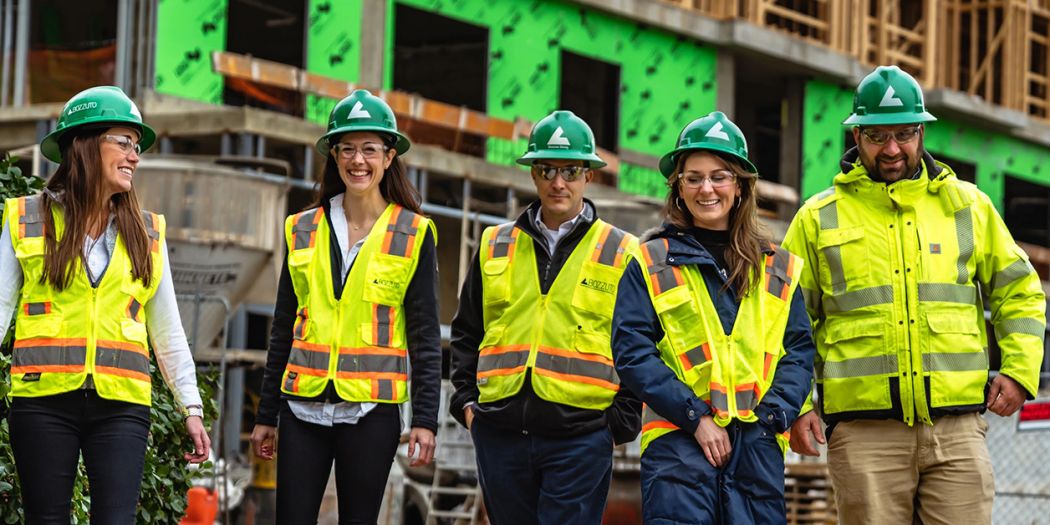Five Bozzuto individuals in safety vests & hard hats posing in front of a construction site.