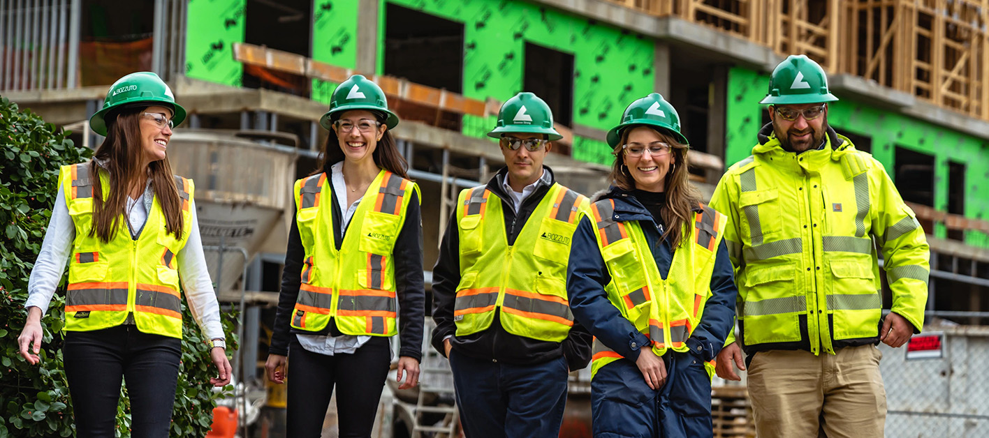 Five Bozzuto individuals in safety vests & hard hats posing in front of a construction site.