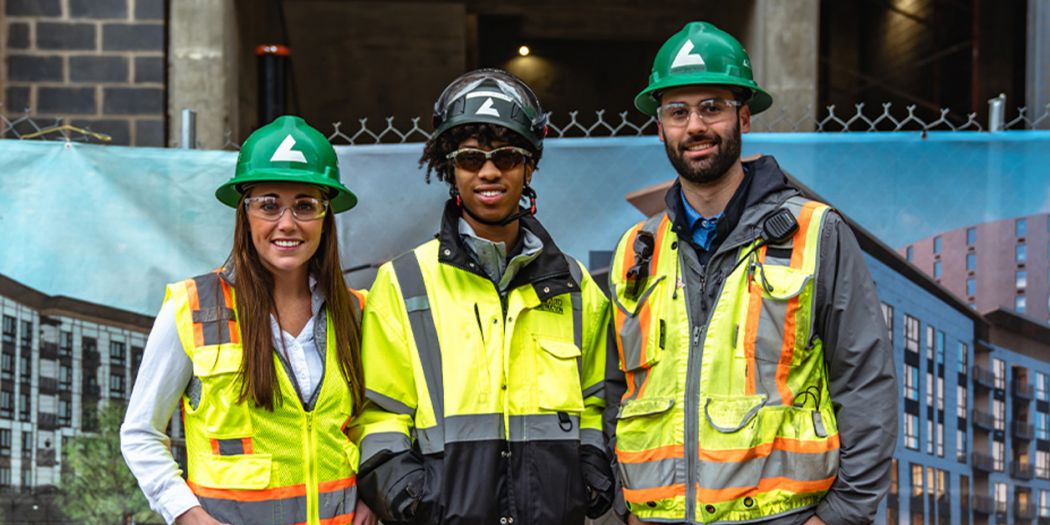 Three Bozzuto individuals in safety vests & hard hats posing in front of a construction site.