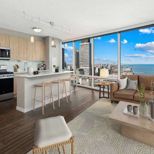 Wide angle of a furnished kitchen and living area, with large windows in the background.