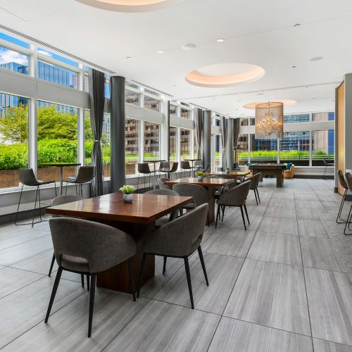 Wide angle of dining tables and bar seating in the resident lounge with large windows and natural light.