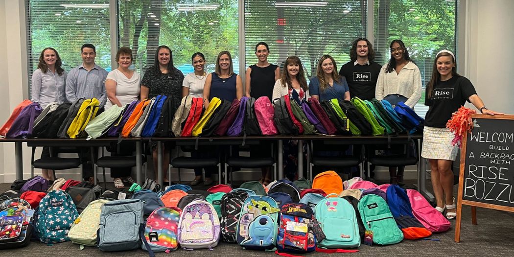A Bozzuto group of individuals standing in front of a stack of backpacks.
