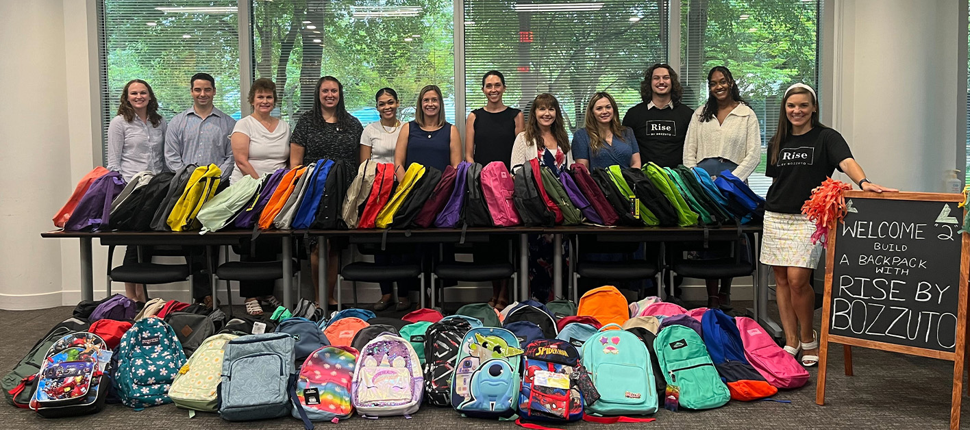 A Bozzuto group of individuals standing in front of a stack of backpacks.