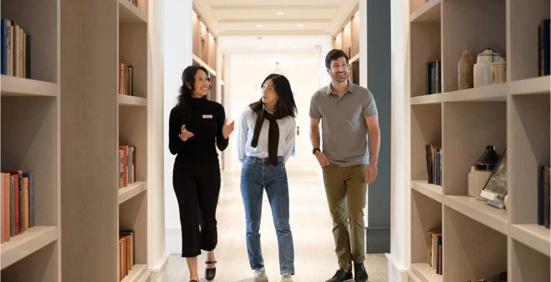 Three people walking in a hallway with bookshelves.